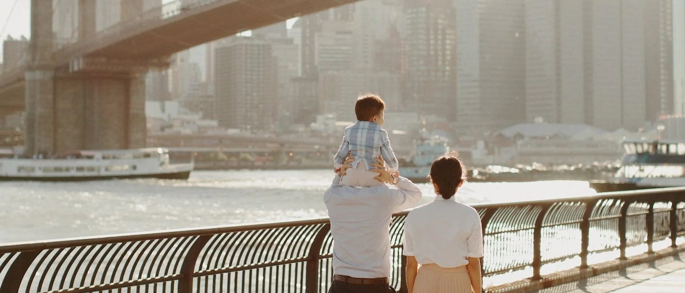 A family of three walks along a waterfront with a city skyline and bridge in the background. The child sits on an adult's shoulders, creating a serene, warm scene.