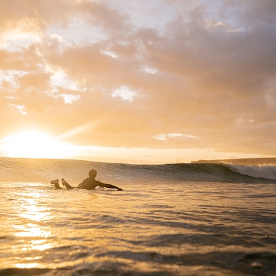 A person lying on a surfboard in the ocean, facing the horizon during sunset. The sky is filled with clouds and the sun is partially hidden, casting a golden glow over the water. 