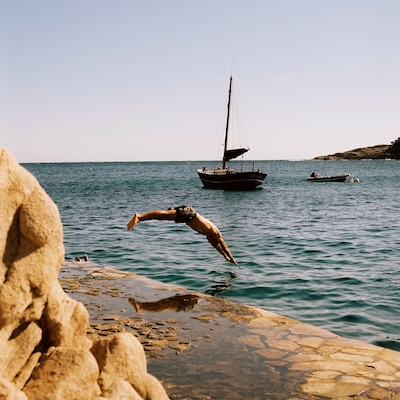 A person diving into the sea from a rocky ledge with two boats in the background under a clear sky.